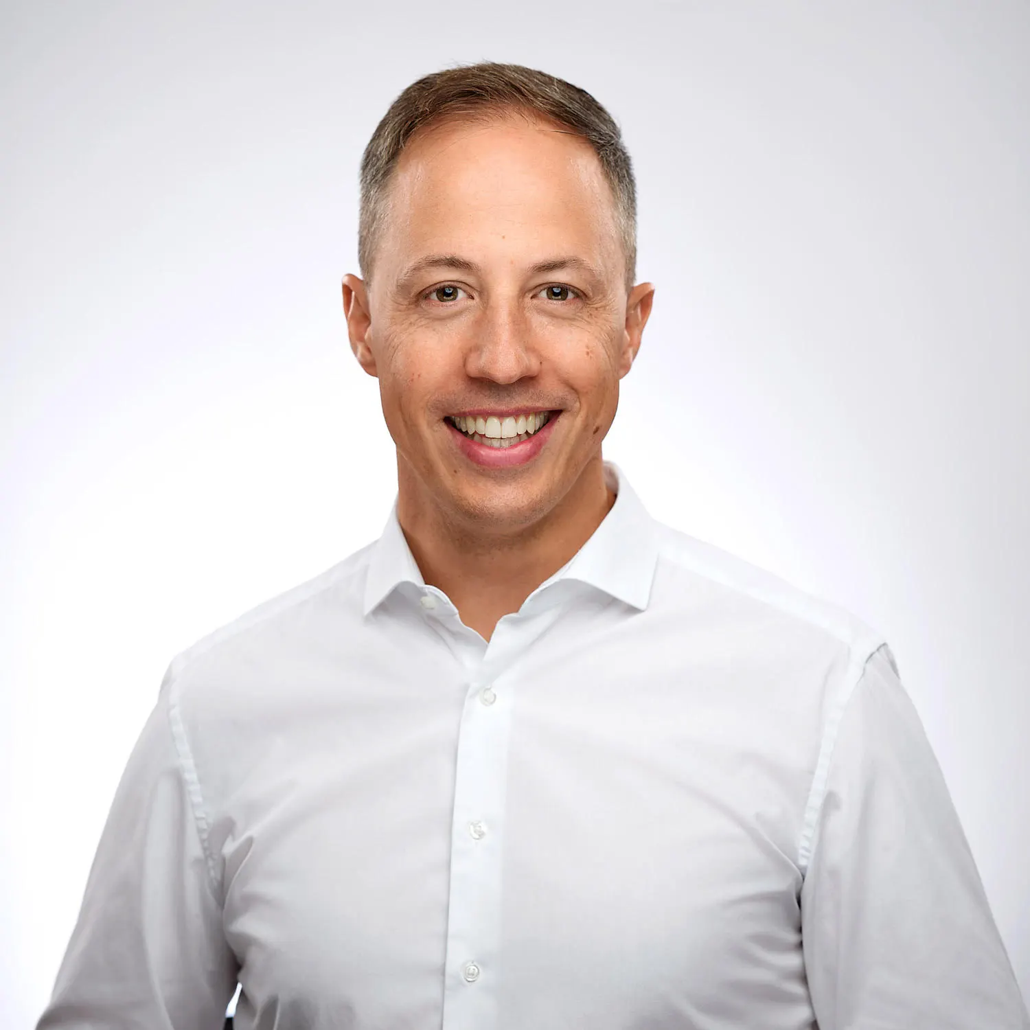 Ein lächelnder Mann mit kurzen Haaren, der eine weiße Hemdbluse trägt. Er blickt direkt in die Kamera vor einem hellgrauen Hintergrund. Sein Name ist Helge Müller-Stephan.