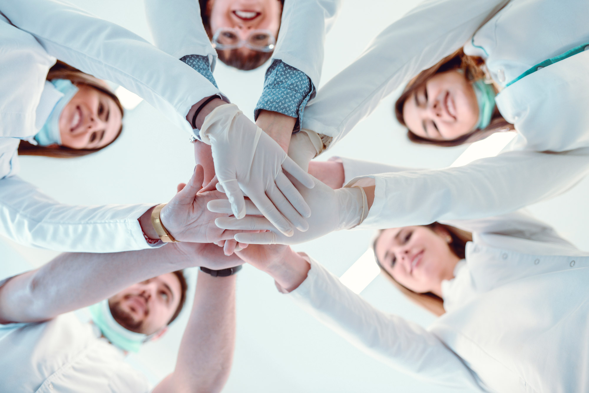 Fünf lachende Mediziner in weißen Kitteln, Handschuhen und Mundschutz legen ihre Hände in der Mitte übereinander.