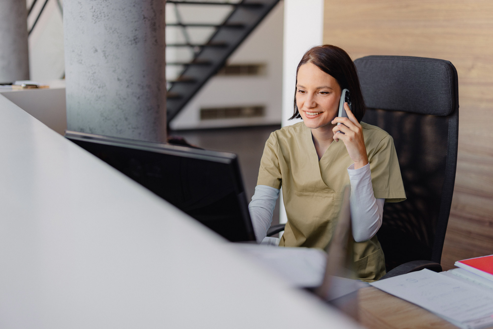 Eine lächelnde Empfangsdame in beiger medizinischer Kleidung sitzt an einem Schreibtisch mit Computer und telefoniert mit einem Mobiltelefon.